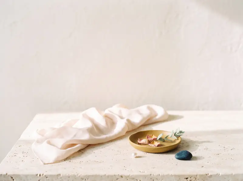 Blush silk draped on cream travertine beside an ochre dish of dried rose petals and sage