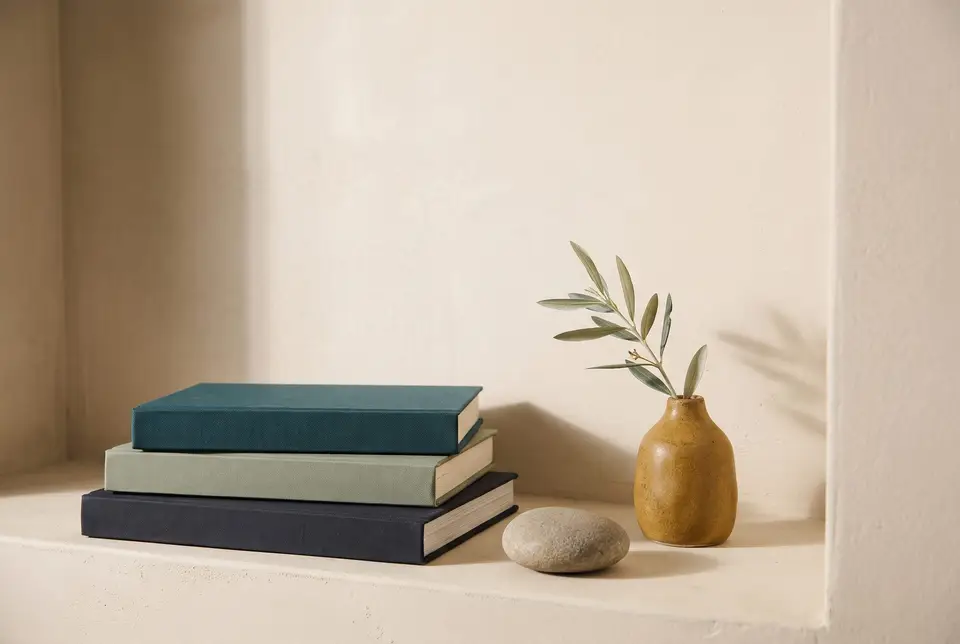Stacked teal, sage and ink cloth books beside a river stone and an ochre vase with an olive sprig on a cream plaster shelf