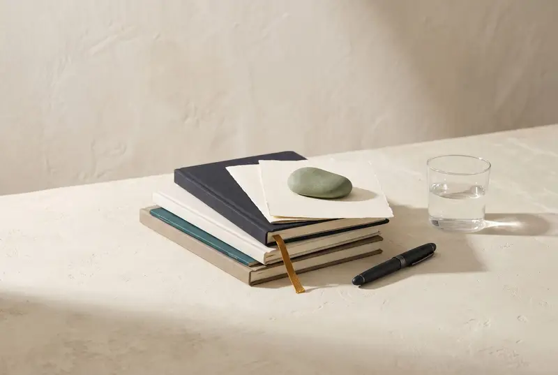 Stack of plain journals with a sage stone paperweight, a fountain pen and a glass of water on a cream surface