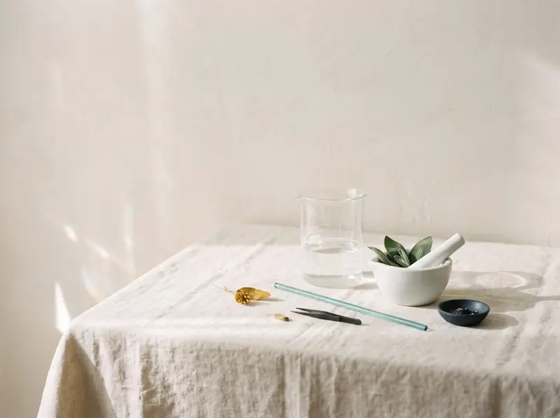 Glass beaker of water and a white mortar with sage leaves on cream linen in soft daylight